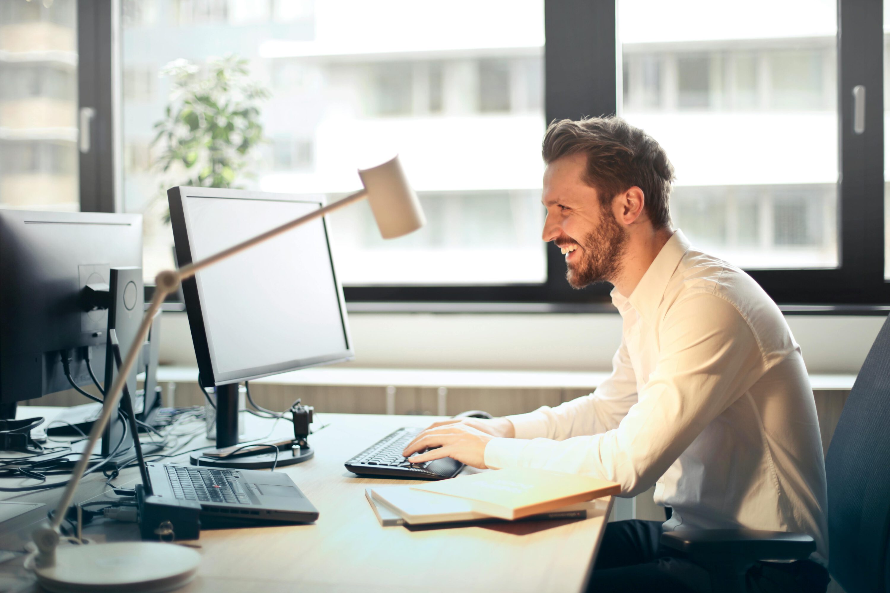 Un homme assis sur une chaise de bureau noire, portant une chemise blanche, souriant tout en travaillant sur un ordinateur de bureau dans un espace lumineux et organisé.