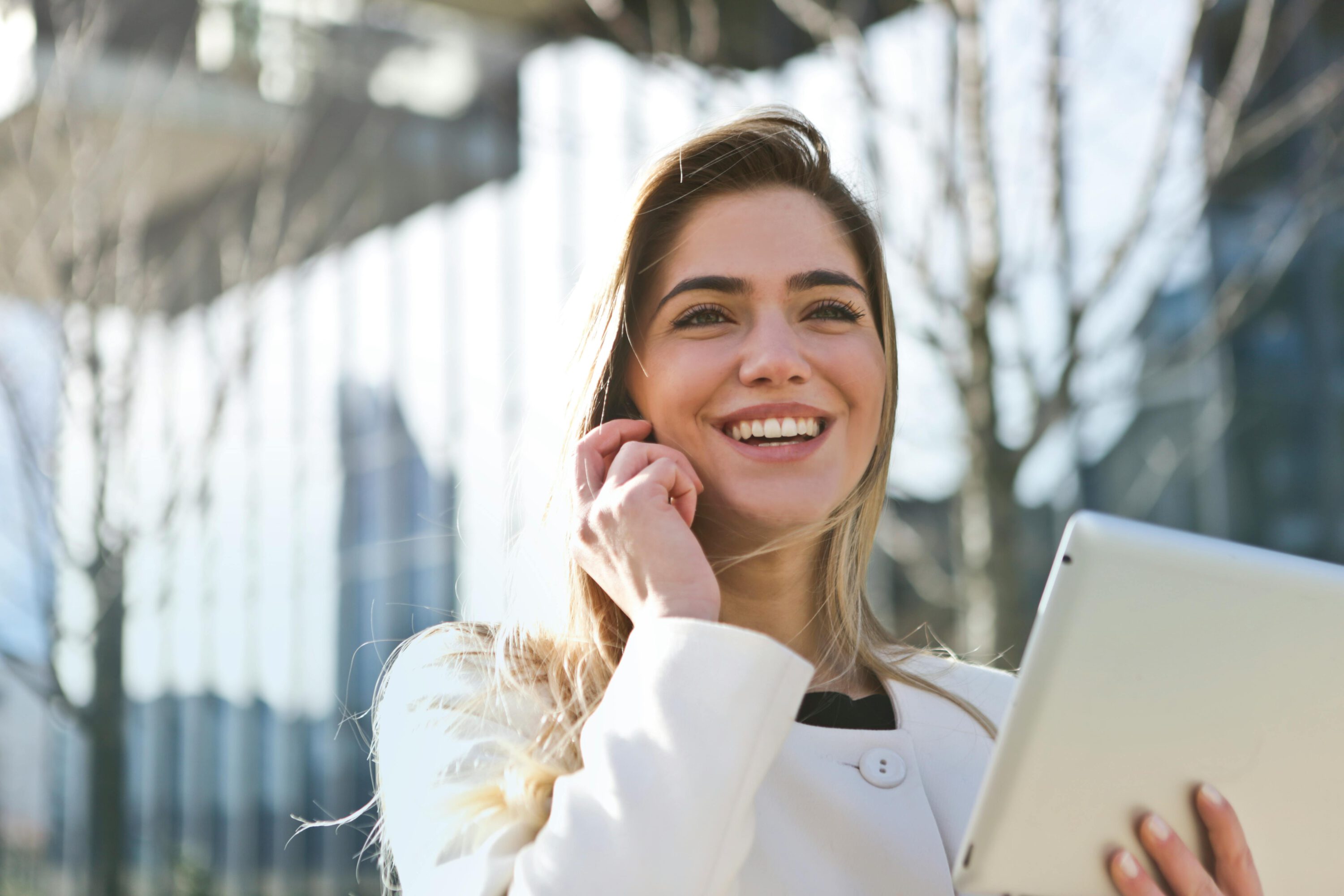 Une femme vêtue d'un blazer blanc, tenant une tablette dans ses mains, avec une expression concentrée, debout dans un environnement professionnel lumineux.